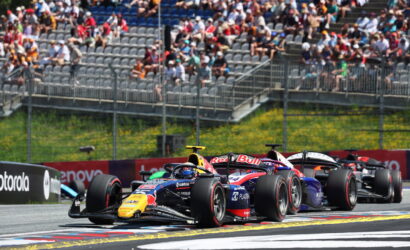 Three Formula 1 race cars cornering on a track as a crowded grandstand watches.