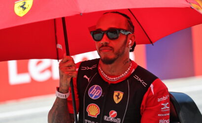 Racing driver in a Ferrari suit standing under a red umbrella with sponsor logos visible on the suit, wearing sunglasses and beads.