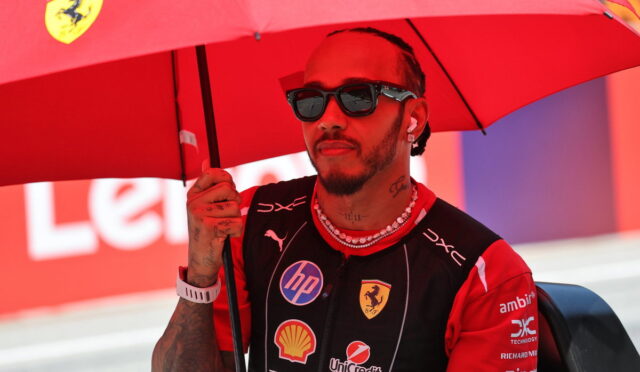 Racing driver in a Ferrari suit standing under a red umbrella with sponsor logos visible on the suit, wearing sunglasses and beads.