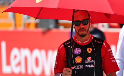 Race car driver in a red Ferrari racing suit holding a red umbrella at a track, wearing dark sunglasses.