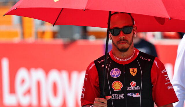 Race car driver in a red Ferrari racing suit holding a red umbrella at a track, wearing dark sunglasses.