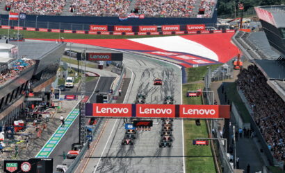 F1 race start on a busy circuit with multiple cars lined up on the grid under large Lenovo banners and a cheering grandstand crowd behind them.