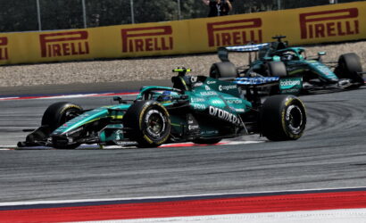 Two teal Formula 1 cars race side by side on a track, with red-and-white curbs and yellow Pirelli barriers in the background, a photographer watching from the fence behind them.