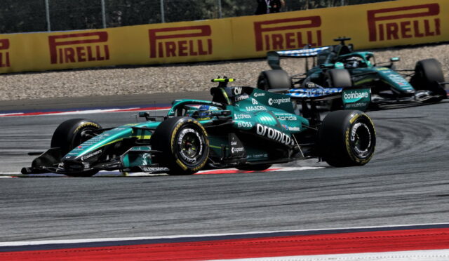 Two teal Formula 1 cars race side by side on a track, with red-and-white curbs and yellow Pirelli barriers in the background, a photographer watching from the fence behind them.