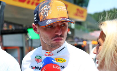 Formula 1 driver in orange and navy cap being interviewed, microphone labeled 'Sport' visible, female interviewer on the right.