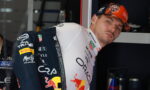 Formula 1 driver in team suit leaning against a garage wall, wearing a Red Bull livery and orange cap.