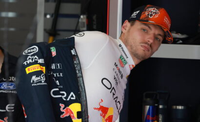 Formula 1 driver in team suit leaning against a garage wall, wearing a Red Bull livery and orange cap.