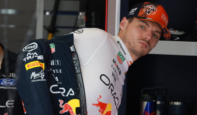 Formula 1 driver in team suit leaning against a garage wall, wearing a Red Bull livery and orange cap.
