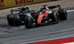 Two Formula 1 cars race side by side through a corner, leaning into the turn on a paved track with Pirelli barriers in the background.