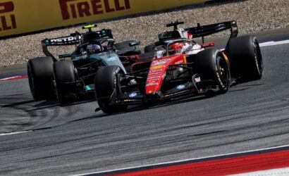 Two Formula 1 cars race side by side through a corner, leaning into the turn on a paved track with Pirelli barriers in the background.