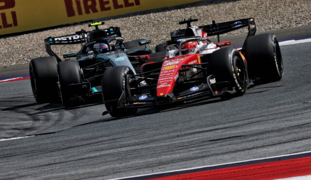Two Formula 1 cars race side by side through a corner, leaning into the turn on a paved track with Pirelli barriers in the background.
