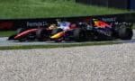 Two Formula 1 cars race side by side on a track, one black/orange Red Bull car and a red‑yellow Oracle‑sponsored car, with grass and barriers in the background.