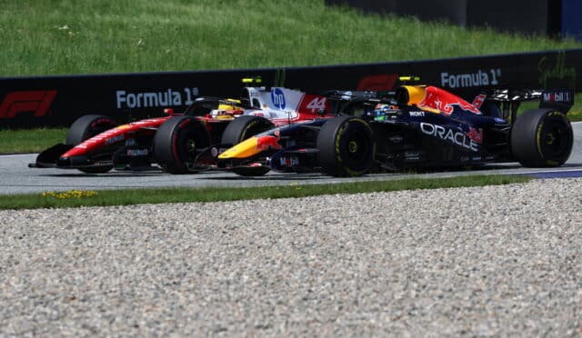 Two Formula 1 cars race side by side on a track, one black/orange Red Bull car and a red‑yellow Oracle‑sponsored car, with grass and barriers in the background.