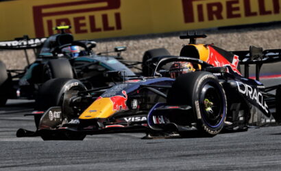 Two Formula 1 cars race side by side on a track, with a Red Bull Oracle-sponsored car in the foreground and a teal Mercedes behind, Pirelli ads in the background.