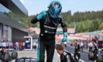 F1 driver in Petronas suit and blue helmet raises a fist in the pit lane, teammates and race cars visible in the background.