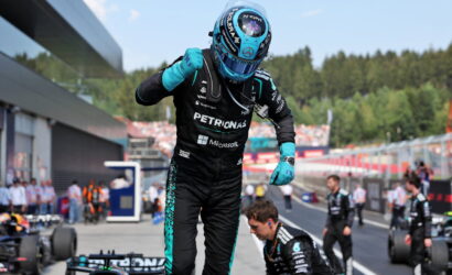 F1 driver in Petronas suit and blue helmet raises a fist in the pit lane, teammates and race cars visible in the background.
