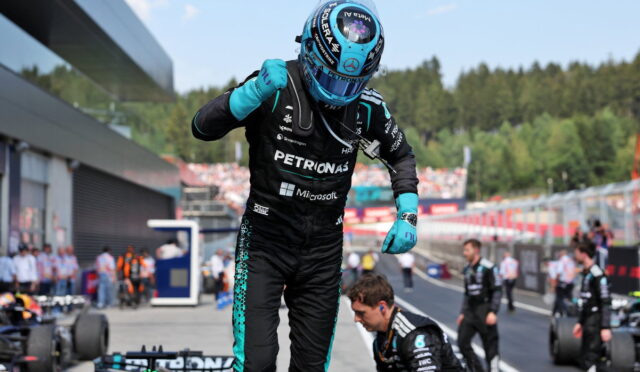 F1 driver in Petronas suit and blue helmet raises a fist in the pit lane, teammates and race cars visible in the background.