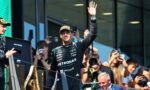 Formula One driver in a Petronas suit waves to fans from a podium with cameras in the background