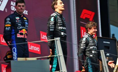 Three race car drivers stand on a podium during a prize ceremony with sponsor banners in the background, on a red stage backdrop.