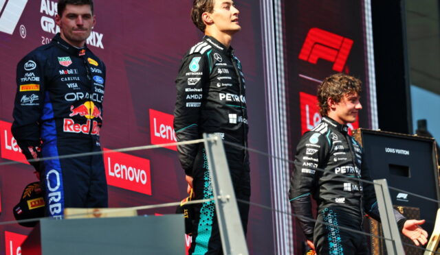 Three race car drivers stand on a podium during a prize ceremony with sponsor banners in the background, on a red stage backdrop.