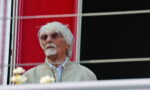 Older man with white hair and goatee wearing round glasses and a light beige jacket, looking to the left.