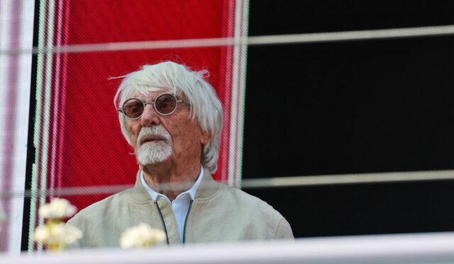 Older man with white hair and goatee wearing round glasses and a light beige jacket, looking to the left.