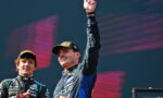 Two race car drivers on a podium, one in the foreground raising his fist in victory. 1st place moment with teammates applauding nearby.