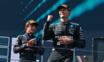 Two Formula 1 drivers on a podium in black Petronas Mercedes suits, one raising a fist in victory.