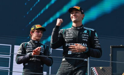 Two Formula 1 drivers on a podium in black Petronas Mercedes suits, one raising a fist in victory.