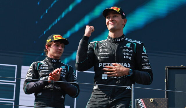 Two Mercedes F1 drivers on a podium celebrate, one fist raised in triumph while the other claps.