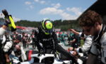 Race car driver in black suit climbs into a white open cockpit while crew members reach in during a pit stop under a blue sky.