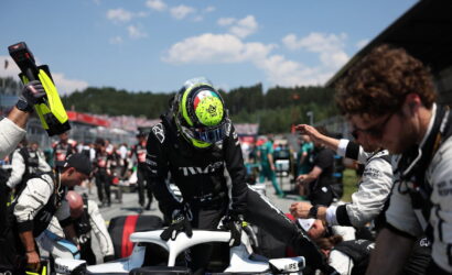 Race car driver in black suit climbs into a white open cockpit while crew members reach in during a pit stop under a blue sky.