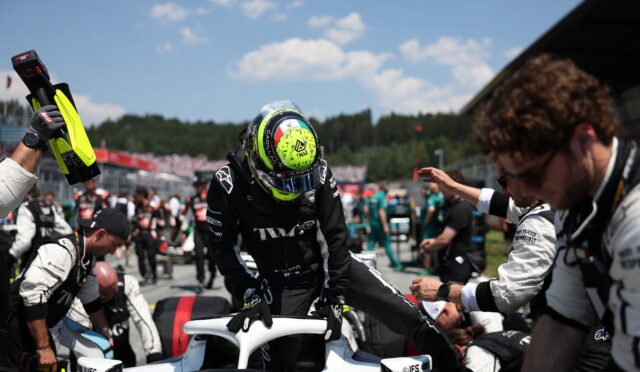 Race car driver in black suit climbs into a white open cockpit while crew members reach in during a pit stop under a blue sky.