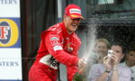 Red-clad race car driver on a podium sprays champagne, smiling as fans with cameras cheer in the background.