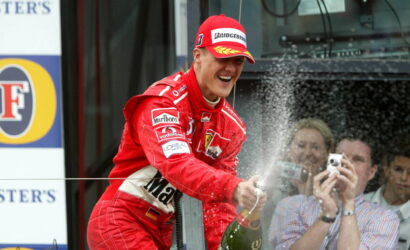 Red-clad race car driver on a podium sprays champagne, smiling as fans with cameras cheer in the background.