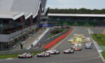 Race cars start a high-speed race on a circuit, with a pit lane on the left and grandstands full of spectators; Rolex signage visible across the track.