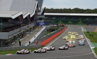 Race cars start a high-speed race on a circuit, with a pit lane on the left and grandstands full of spectators; Rolex signage visible across the track.