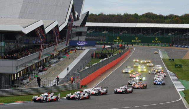 Race cars start a high-speed race on a circuit, with a pit lane on the left and grandstands full of spectators; Rolex signage visible across the track.