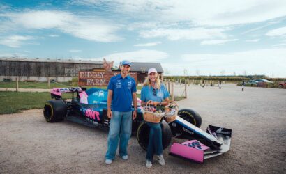 Two people stand beside a pink and blue race car on a gravel lot, with a wooden 'DIDDLY FARM' sign behind them.