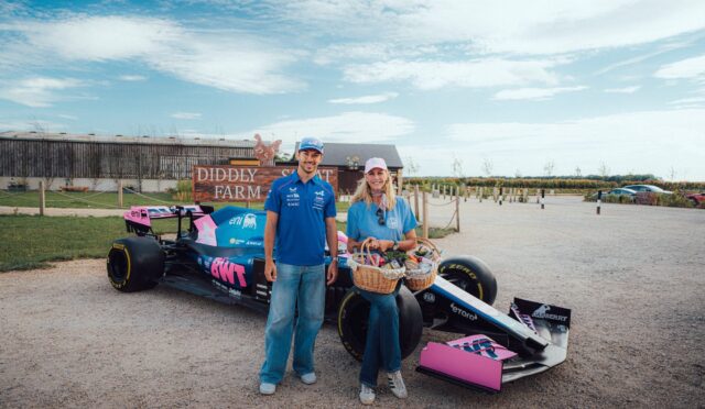 Two people stand beside a pink and blue race car on a gravel lot, with a wooden 'DIDDLY FARM' sign behind them.