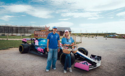 Man and woman in blue shirts pose beside a blue Formula-style race car on a gravel lot with a Diddly Farm sign in the background, holding a gift basket.