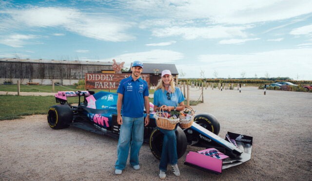 Man and woman in blue shirts pose beside a blue Formula-style race car on a gravel lot with a Diddly Farm sign in the background, holding a gift basket.