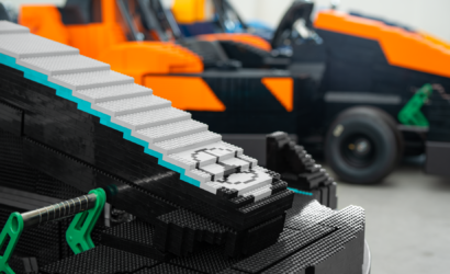 Close-up of a LEGO staircase with grey steps and teal accents on a black LEGO base, blurred orange machinery in the background.