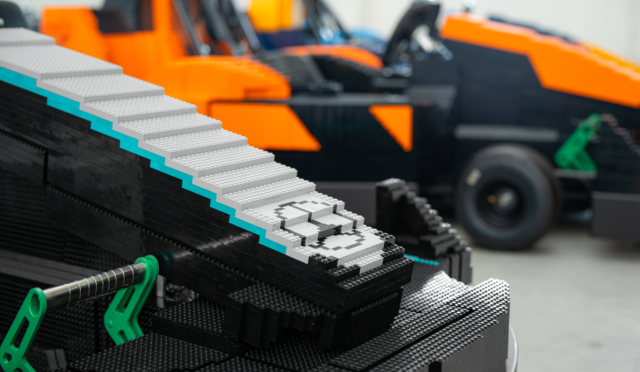 Close-up of a LEGO staircase with grey steps and teal accents on a black LEGO base, blurred orange machinery in the background.