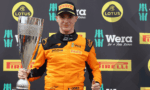 Young race driver in orange suit holding a tall trophy at a sponsor-filled backdrop (Lotus, Pirelli, Wera).