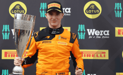Young race driver in orange suit holding a tall trophy at a sponsor-filled backdrop (Lotus, Pirelli, Wera).