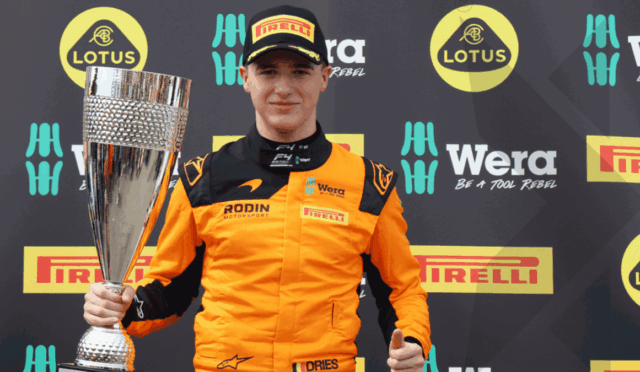 Young race driver in orange suit holding a tall trophy at a sponsor-filled backdrop (Lotus, Pirelli, Wera).