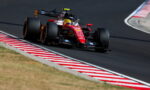 Red Formula 1 race car taking a tight turn on a race circuit, red-and-white striped curbs visible on the inside edge.