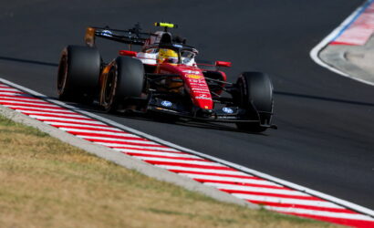 Red Formula 1 race car taking a tight turn on a race circuit, red-and-white striped curbs visible on the inside edge.
