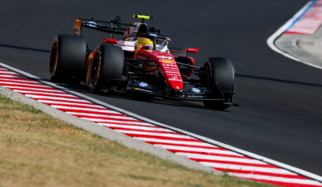 Red Formula 1 race car taking a tight turn on a race circuit, red-and-white striped curbs visible on the inside edge.